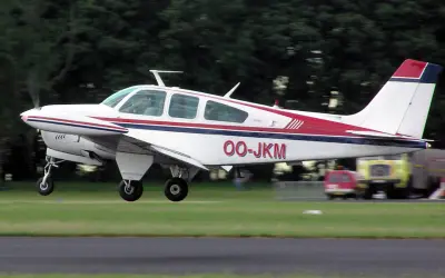 Eine weiße Beechcraft Bonanza mit roten und blauen Streifen hebt vom Gras ab.