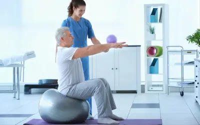Starszy mężczyzna ćwiczy z fizjoterapeutką na piłce, co może być częścią rehabilitacji.