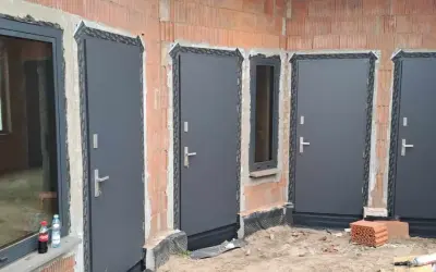 Nowe, szare drzwi zewnętrzne i okno w budowie. Widać ceglane ściany i materiały budowlane.