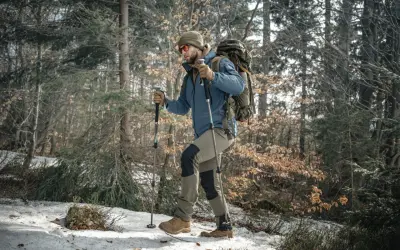 Mężczyzna w czapce i okularach przeciwsłonecznych idzie przez zaśnieżony las, wspierając się na kijach trekkingowych.