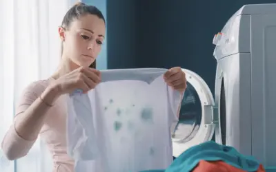 Zmartwiona kobieta trzyma białą koszulkę z zielonymi plamami, widoczne odbarwione ubrania po praniu.