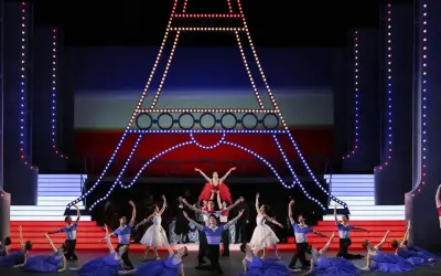 Tancerze baletowi na scenie z miniaturą Wieży Eiffla. W tle czerwono-niebieska scenografia, przypominająca flagę Francji. W powietrzu unosi się aria.