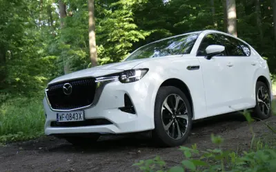 Biała Mazda CX-60 zaparkowana na leśnej ścieżce, otoczona zielenią drzew i traw.