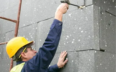 Budowlaniec w żółtym kasku i kamizelce nakłada piankę montażową na styropian. Grubość styropianu jest kluczowa dla izolacji.