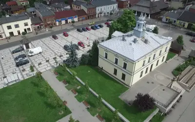 Sławków rynek z lotu ptaka: żółty budynek ratusza, brukowany plac z samochodami i zielony park z ławkami.