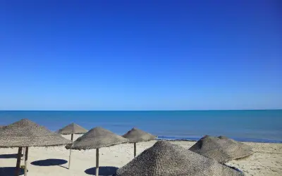 Słoneczna plaża na Djerbie w kwietniu. Puste parasole z trawy na piasku, turkusowe morze i bezchmurne niebo.