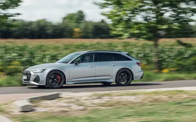 Srebrne Audi RS6 Avant pędzi po wiejskiej drodze, otoczone zielenią pól i drzew.