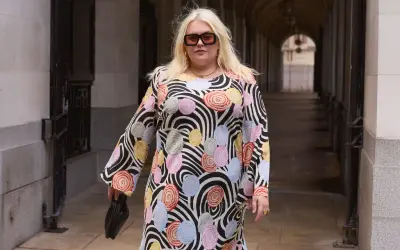 Kobieta w kolorowej tunice w kwiaty i paski, w okularach, trzyma czarną torebkę.