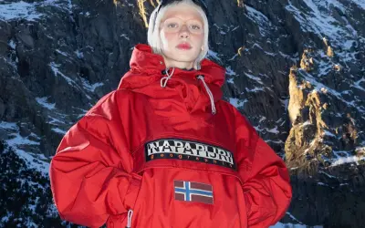 Modelka w czerwonej kurtce z logo Napapijri Geographic i flagą Norwegii na tle ośnieżonych gór. Napapijri co to za firma? To marka odzieży outdoorowej.