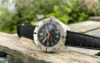 Reloj Sandoz Geneve Typhoon con correa negra, sobre madera rústica y fondo verde.