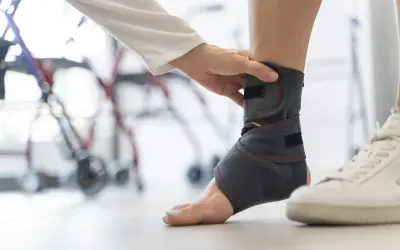 Dłoń zakłada ściągacz na kostkę, by zapewnić jej stabilizację. W tle widać rehabilitacyjny sprzęt.