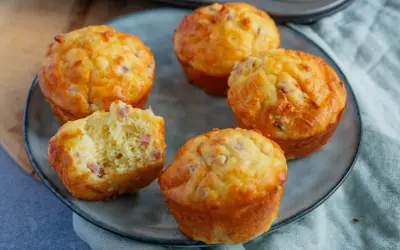 Fünf herzhafte Muffins mit Speck und Käse auf einem grauen Teller. Einer ist angeschnitten und zeigt die saftige Krume.
