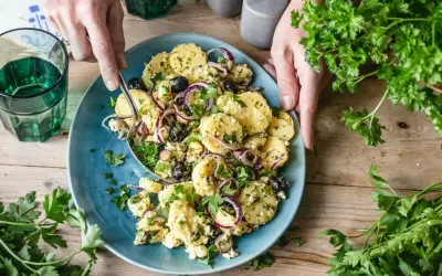 Ein köstlicher mediterraner Kartoffelsalat mit Oliven, roten Zwiebeln und Feta auf einem blauen Teller, garniert mit frischer Petersilie.