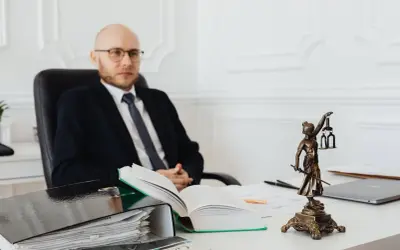 Prawnik w garniturze siedzi przy biurku, obok niego stos dokumentów i Statua Temidy. Zastanawiasz się, ile kosztuje notariusz przy zakupie mieszkania?
