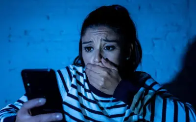 Przerażona kobieta trzyma telefon, jej dłoń zakrywa usta. Czyta wiadomość, która jest ewidentnym szantażem.