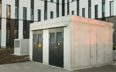 Betonowa stacja transformatorowa z dwoma czarnymi drzwiami i żółtymi znakami ostrzegawczymi. W tle nowoczesny budynek z białymi i ciemnymi panelami.