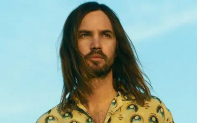 Kevin Parker w koszuli z motywem oczu, z rozwianymi włosami na tle błękitnego nieba.