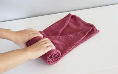 Dłonie zwijają różowy sweter, pokazując, jak składać swetry, by zajmowały mniej miejsca.