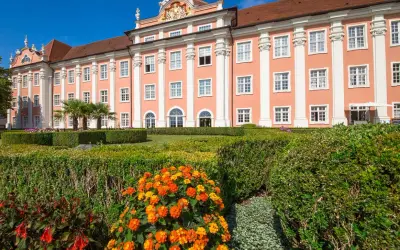 Das Meersburg Neues Schloss erstrahlt in Rosa vor blauem Himmel. Ein gepflegter Garten mit bunten Blumen und Palmen lädt zum Verweilen ein.