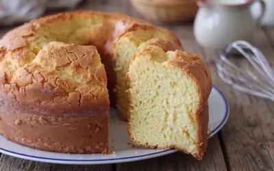Una torta alta, soffice e dorata, con una fetta tagliata che rivela la sua morbidezza. Sullo sfondo, uova e latte, ingredienti per un dolce fatto in casa.