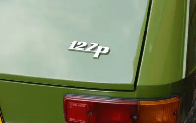 Chromowana plakietka "127p" na zielonej karoserii klasycznego Fiata 127. Widoczny fragment tylnej lampy.