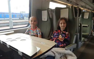 Dzieci siedzą w przedziale dla osoby z dzieckiem do lat 6 Intercity. Chłopiec w kolorowym T-shircie i dziewczynka w kurtce z kwiatowym wzorem.