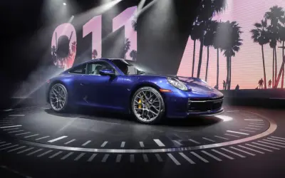 Un Porsche 911 992 azul metálico, con pinzas de freno amarillas, se exhibe sobre un escenario circular con un fondo de palmeras y el número 011.