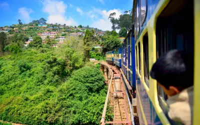 Ett blått och gult tåg i Indien färdas längs en bergssluttning täckt av frodig grönska och färgglada hus.