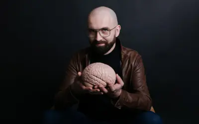 Łysy mężczyzna w okularach i brązowej kurtce trzyma w dłoniach model mózgu.