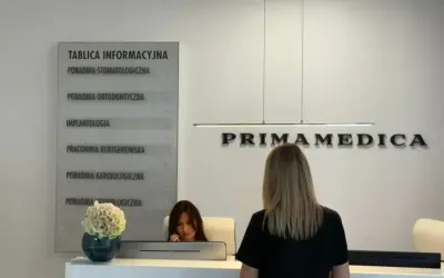 Recepcja kliniki PRIMAMEDICA z tablicą informacyjną i pracownicą.