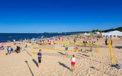 Plaża Brzeźno tętni życiem. Ludzie grają w siatkówkę plażową, opalają się i spacerują. W tle widać morze i namioty.