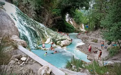 Cascata bianca e pozze azzurre alle terme San Filippo, con persone che si rilassano nell'acqua termale.