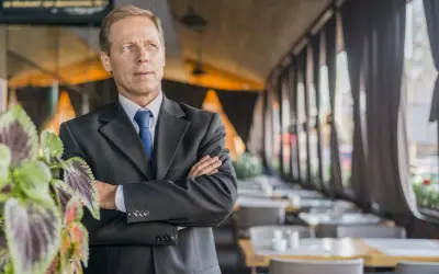 Uomo d'affari con braccia conserte, un chief people officer che guarda lontano, con piante verdi in primo piano e un ristorante sullo sfondo.