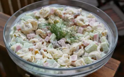Pyszna sałatka z tortellini, szynką i ogórkiem, udekorowana gałązką koperku. Idealna na letnie przyjęcie.