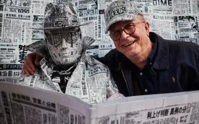 Tomasz Tomaszewski w kapeluszu i płaszczu z gazet. Obok uśmiechnięty mężczyzna w okularach.