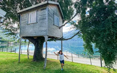 Bambina corre verso una casa sull'albero in legno, con un lago e montagne sullo sfondo.