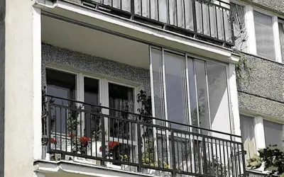 Zabudowa balkonu w bloku z przeszklonymi drzwiami i metalową balustradą. Na balkonie stoją doniczki z kwiatami.