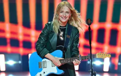 Uśmiechnięta polska piosenkarka z blond włosami gra na niebieskiej gitarze elektrycznej na tle pomarańczowych świateł.