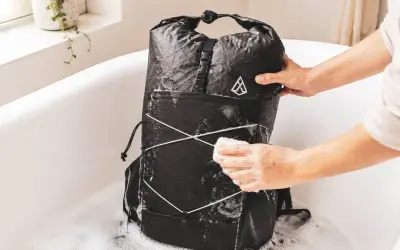 Person reinigt einen schwarzen Rucksack in einer Badewanne mit Schaum. Der Rucksack wird mit einem Schwamm abgerieben, um ihn zu waschen.