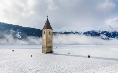 Torre sommersa nel lago di Resia, circondata da neve e montagne nebbiose. Alcune persone passeggiano sul ghiaccio.