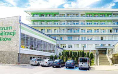 Sanatorium z rehabilitacją kręgosłupa dla rolników. Nowoczesny budynek z zielonymi elementami fasady, otoczony drzewami i parkingiem.