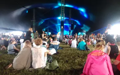 Widownia siedzi na trawie pod namiotem, czekając na koncert. Niebieskie światła oświetlają scenę, tworząc atmosferę festiwalu Open'er.