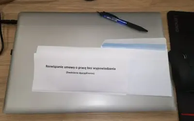 Na biurku leży laptop, na nim dokument z napisem "Rozwiązanie umowy o pracę bez wypowiedzenia (zwolnienie dyscyplinarne)". Obok długopis i myszka.