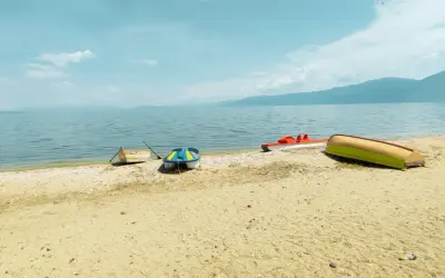 Spokojny brzeg Jeziora Ochrydzkiego w Albanii. Kilka łódek, w tym kolorowa łódka i czerwona rowerowa, odpoczywa na piaszczystej plaży.