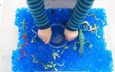 Kinderhände erkunden eine Kiste voller blauer Wasserperlen mit Spielzeug-Echsen und Schlangen. Tolle Wasserperlen Ideen für sensorisches Spiel!