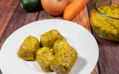 Cinque cubetti dorati di dado vegetale fatto in casa, preparati con carote, zucchine e cipolle, su un piatto bianco.