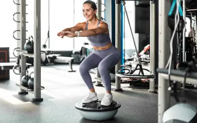 Frau macht Kniebeugen auf einem Bosu-Ball, trägt Sportkleidung und weiße Sneaker. Sie trainiert ohne schuhe auf dem instabilen Untergrund.