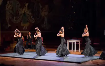 Cuatro bailarinas de flamenco, con vestidos negros voluminosos, ejecutan pasos apasionados. Un hombre aplaude al fondo.