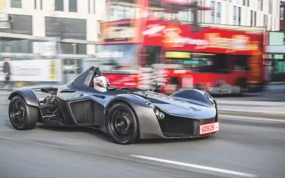 Niesamowity BAC Mono pędzi ulicą, mijając rozmazany czerwony autobus i budynki.