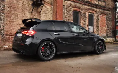Czarny Mercedes-AMG A45s z masywnym spojlerem i czerwonymi zaciskami hamulcowymi stoi na mokrej nawierzchni przed starą ceglaną kamienicą.